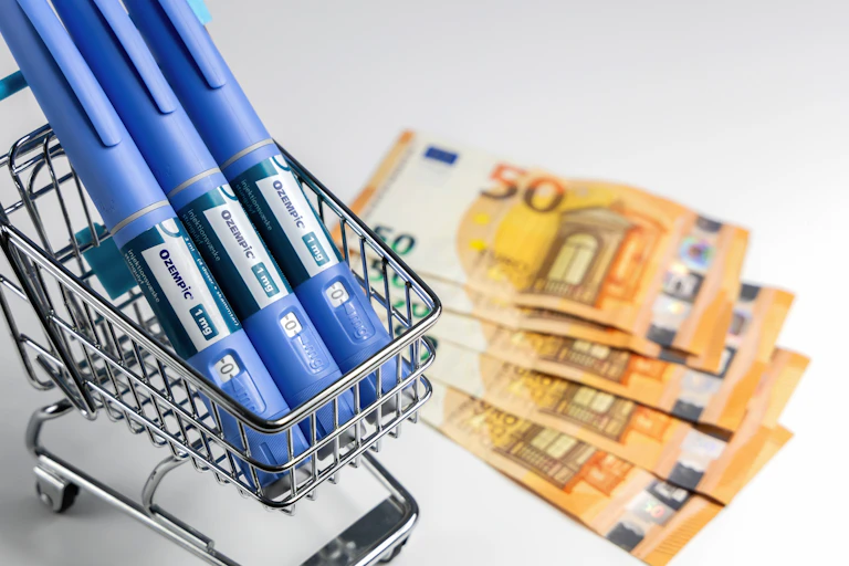 Three Ozempic pens in a miniature supermarket trolley with cash notes next to them.