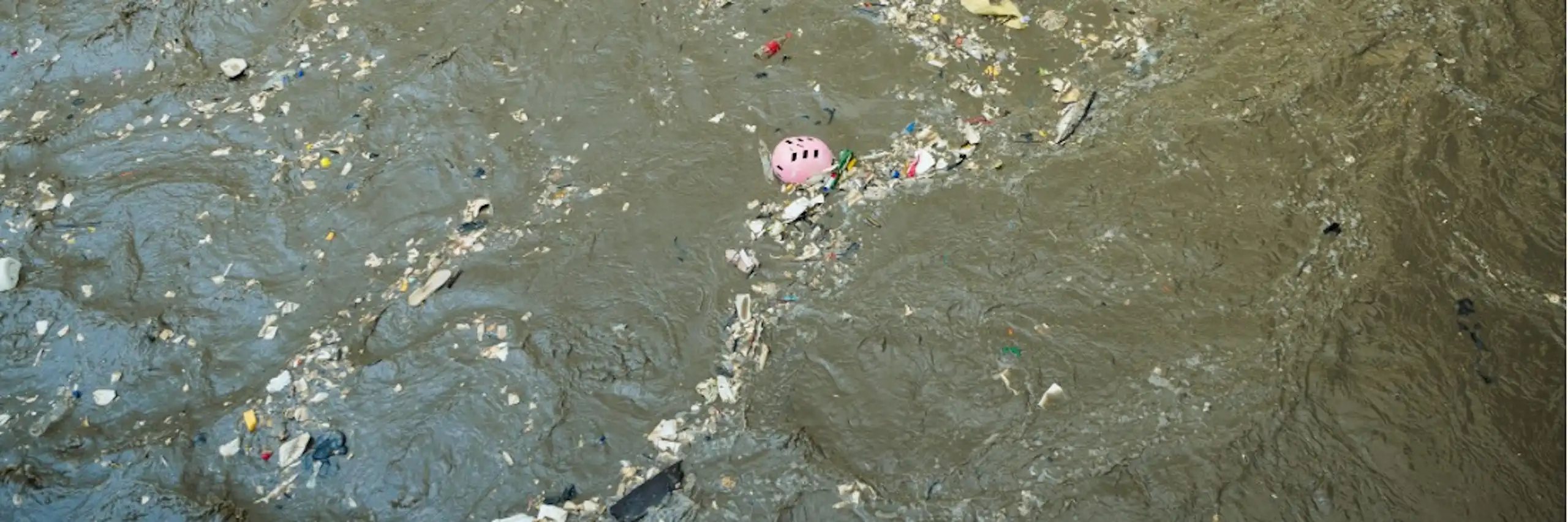 Vue de déchets plastiques (casque de vélo, bouteilles vides, emballages) flottant à la surface de l’eau.