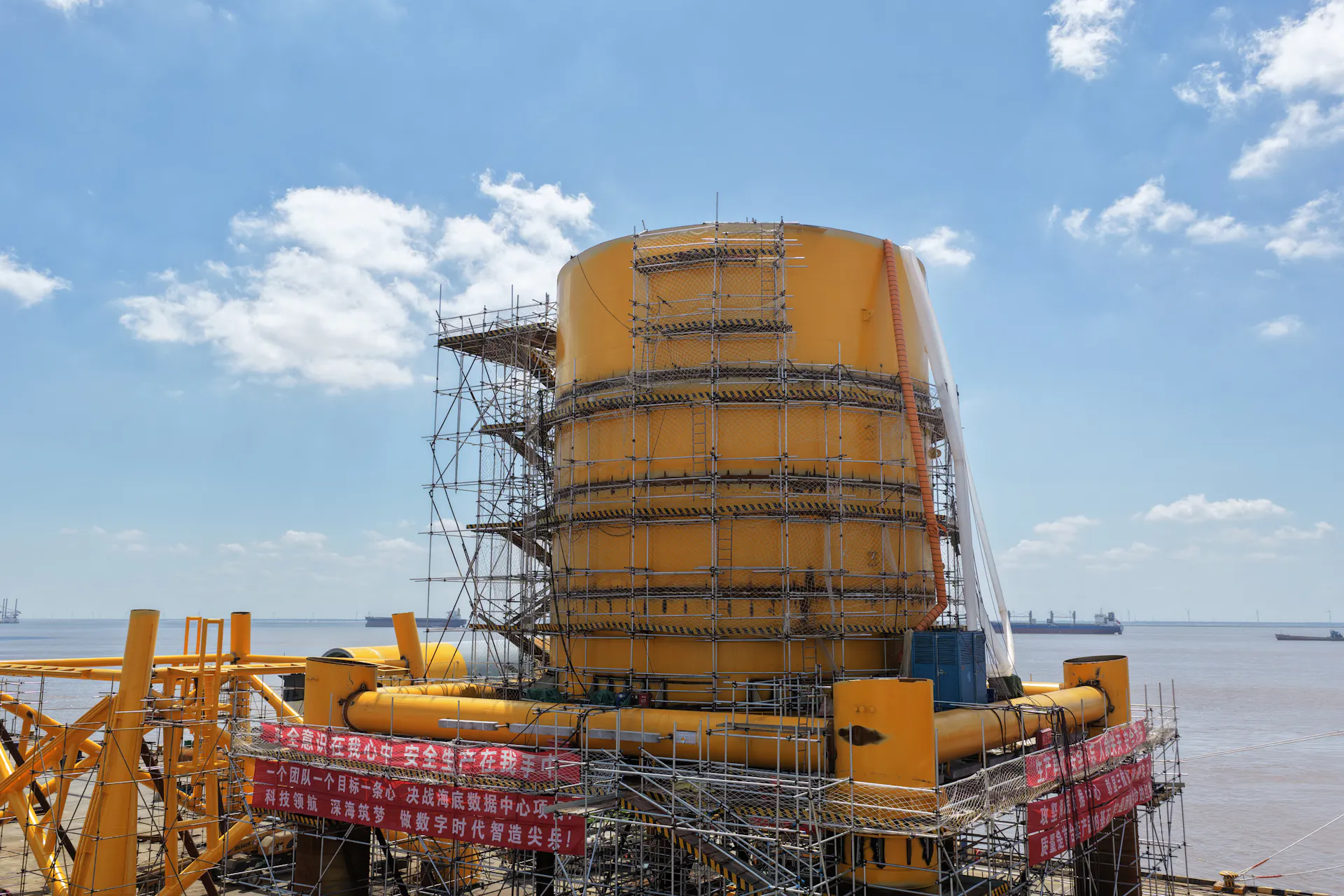 A large yellow cylinder surrounded by scaffolding sits on a platform next to a body of water.