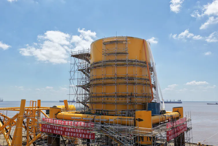 A large yellow cylinder surrounded by scaffolding sits on a platform next to a body of water.