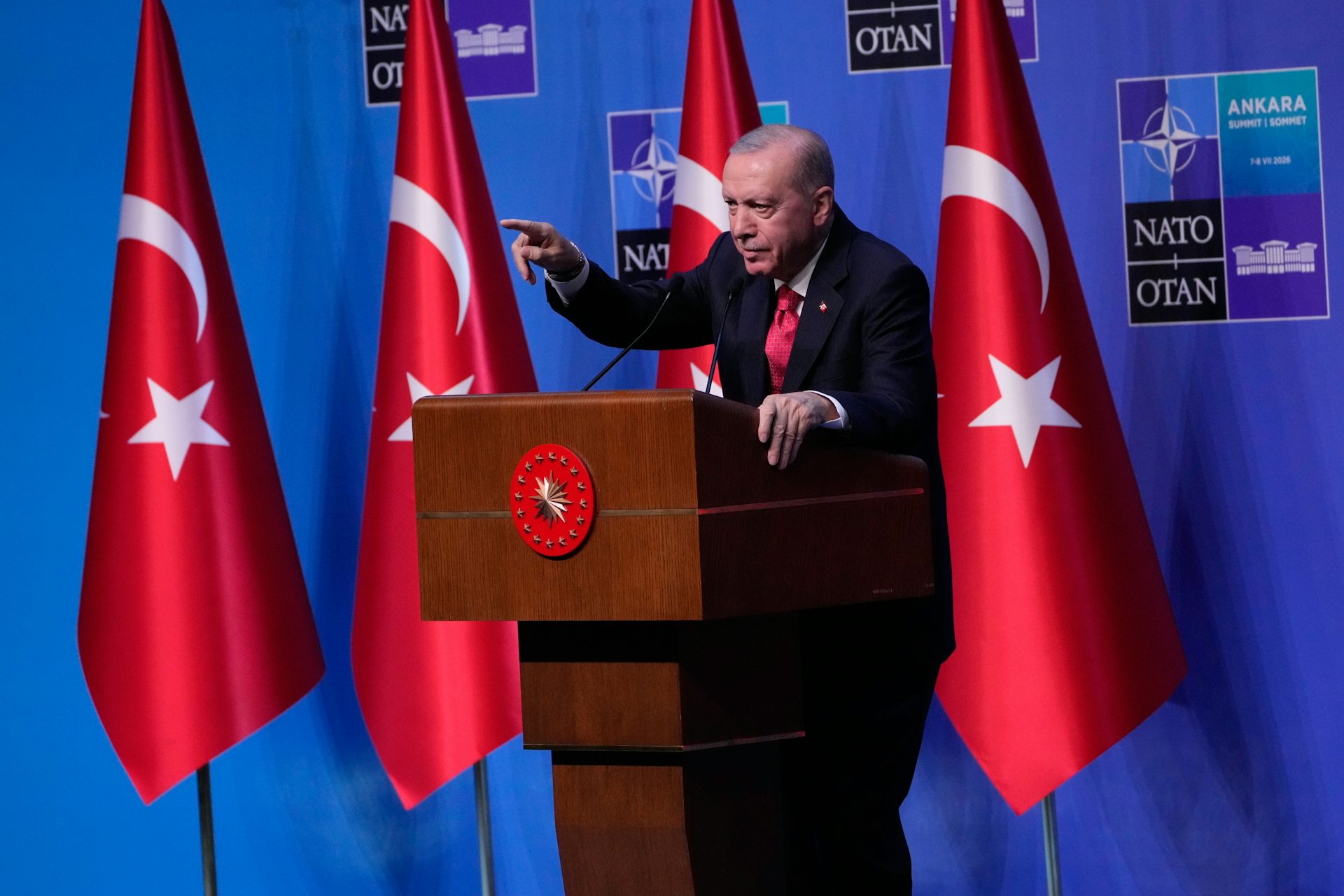 A man in a dark suit behind a podium with red flags behind him.