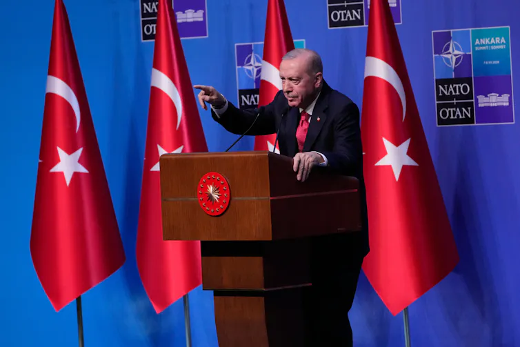 A man in a dark suit behind a podium with red flags behind him.
