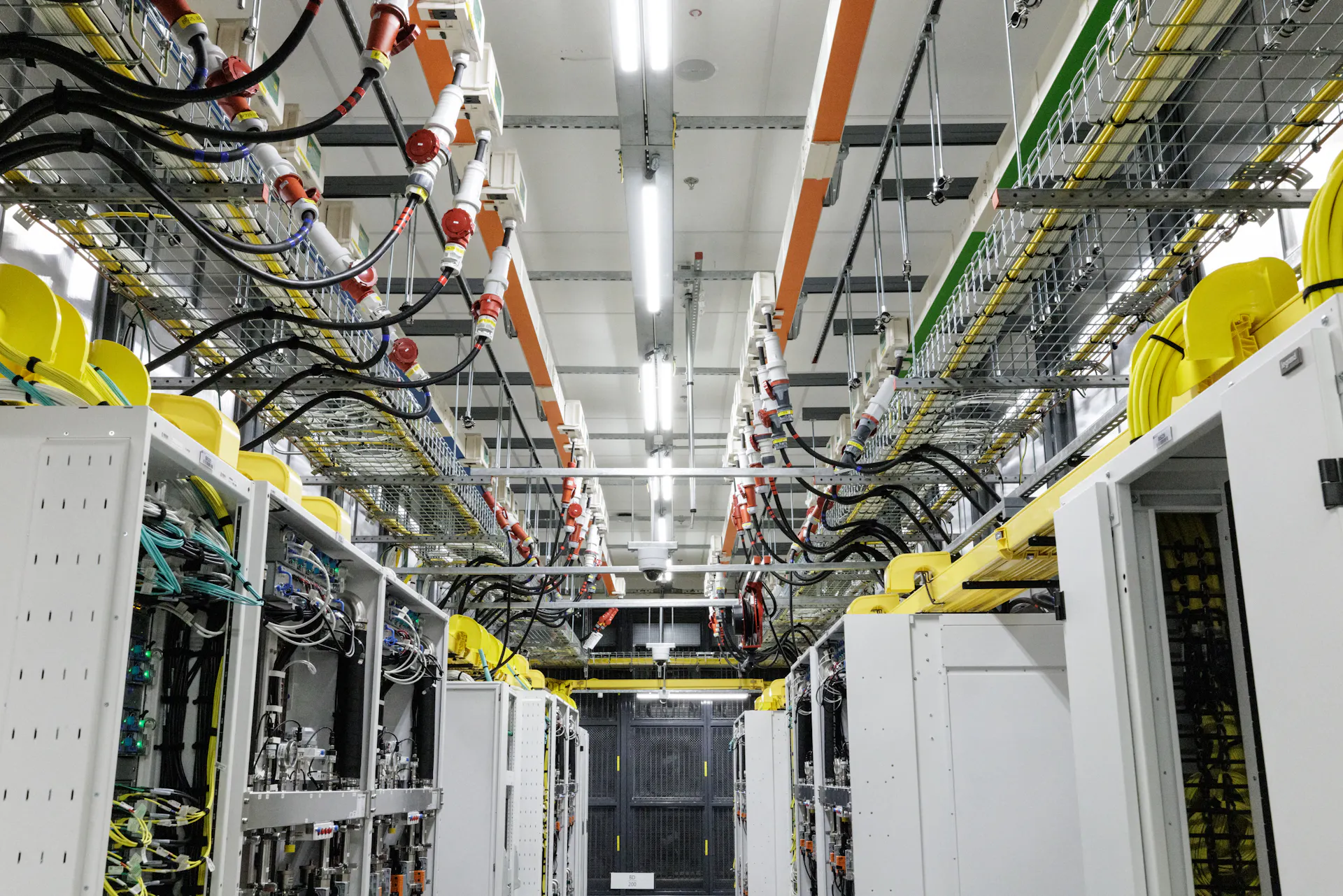 A view of an industrial room with metal boxes in rows on the floor and pipes and tubes running overhead.