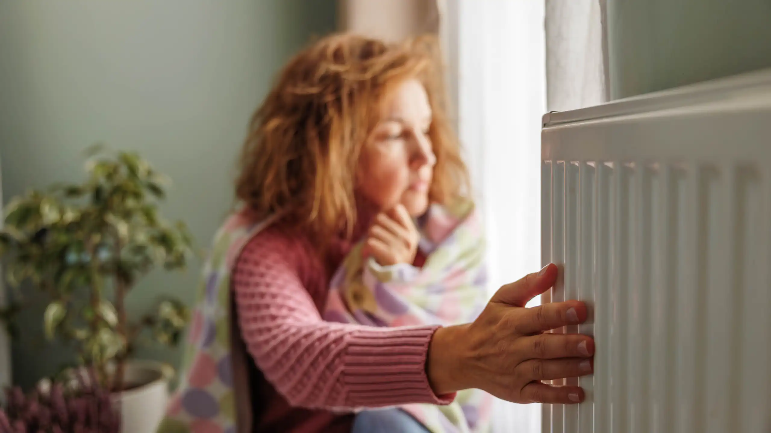 A person wrapped in a blanket sits next to a radiator, with her hand on the heating element.