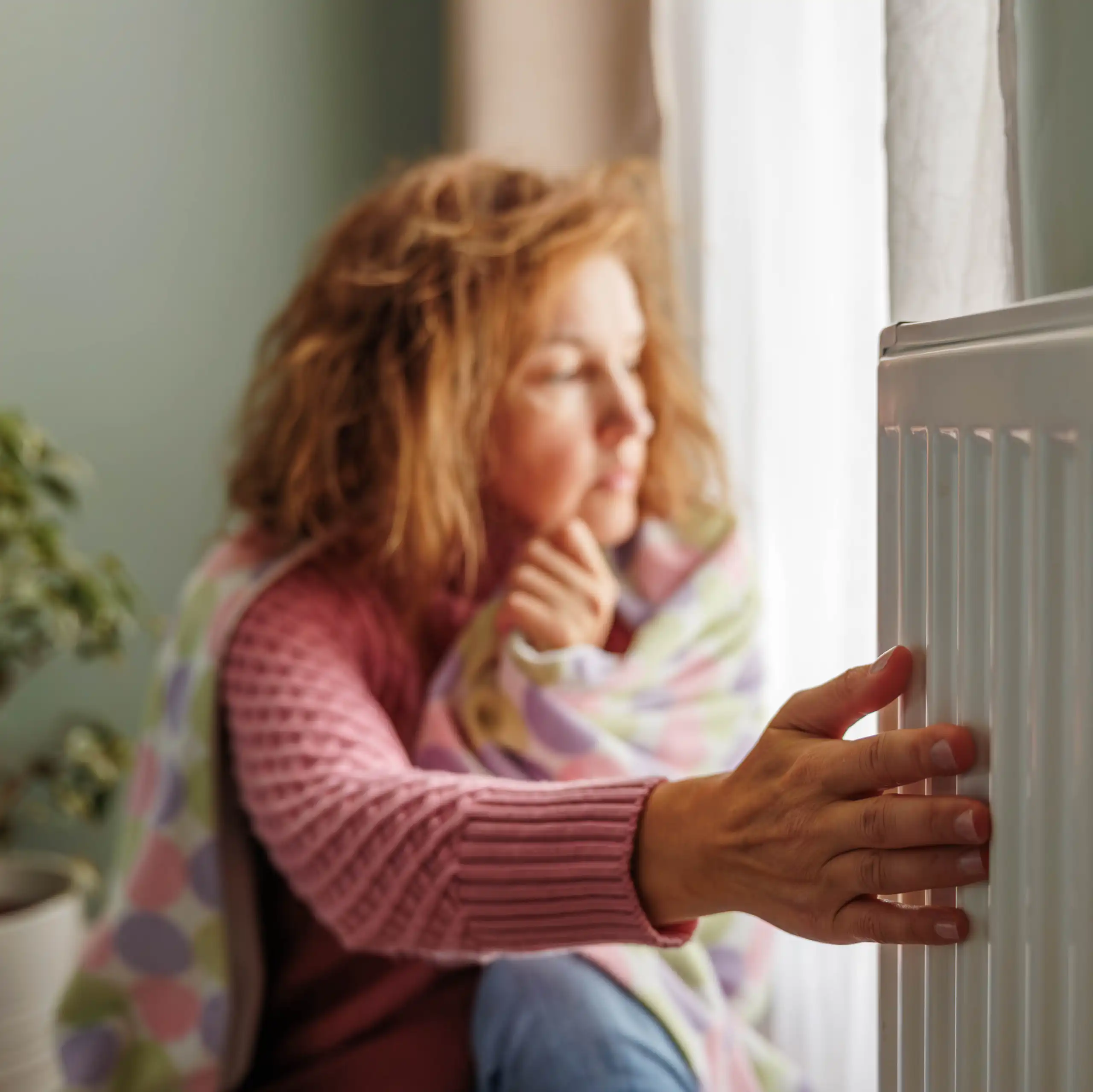 A person wrapped in a blanket sits next to a radiator, with her hand on the heating element.