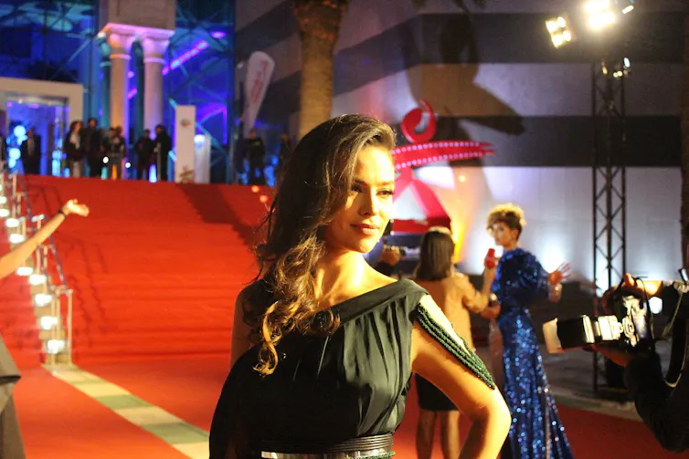 A vast red carpet with people mingling at the top of the stairs it goes up. In the foreground a smartly dressed woman poses for photographers.