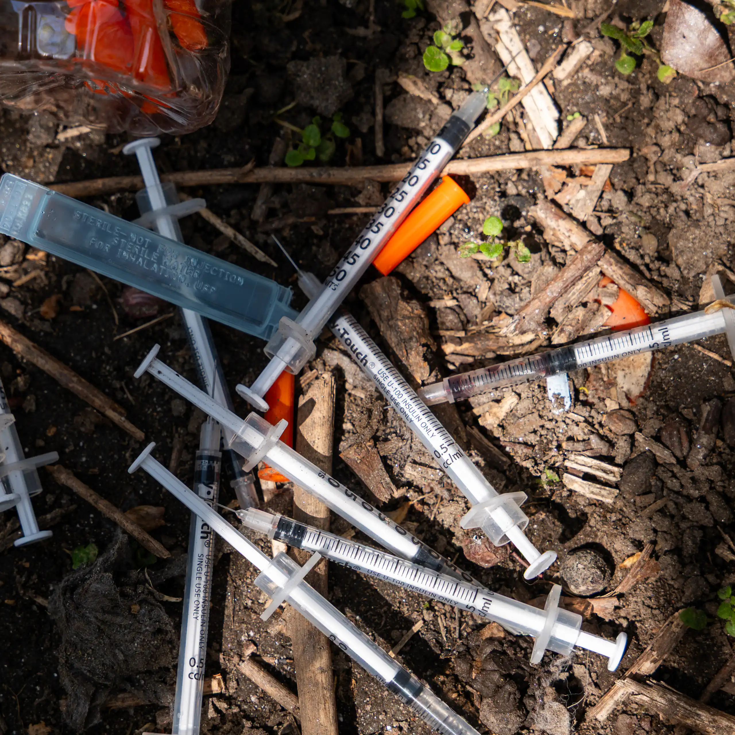 Used needles with orange caps lie in the dirt.