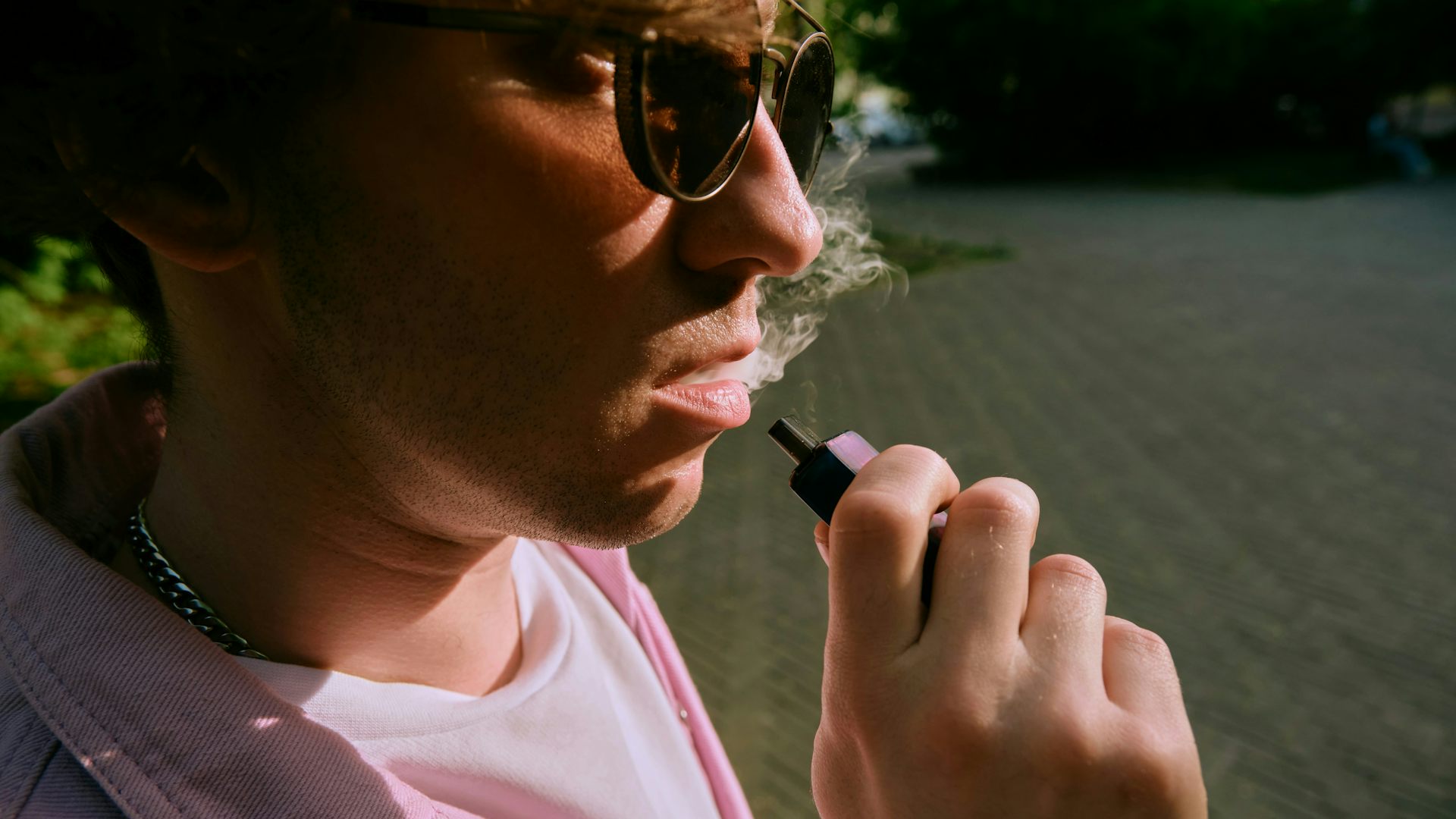 Close-up photo of a young man in a pink button-up shirt and sunglasses vaping