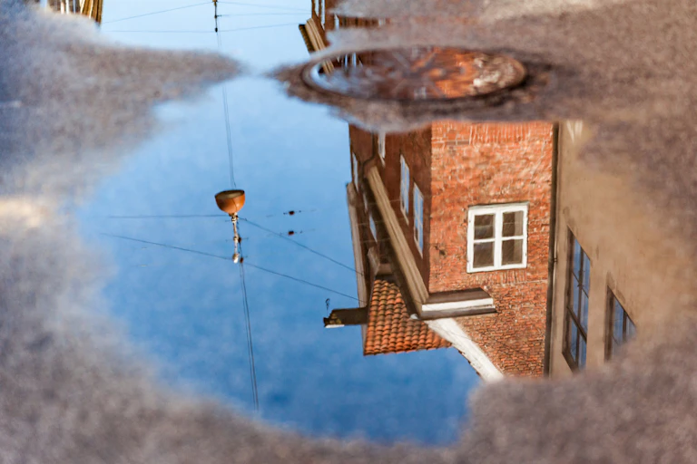 The reflection of a brick house is seen in a small puddle on the road.