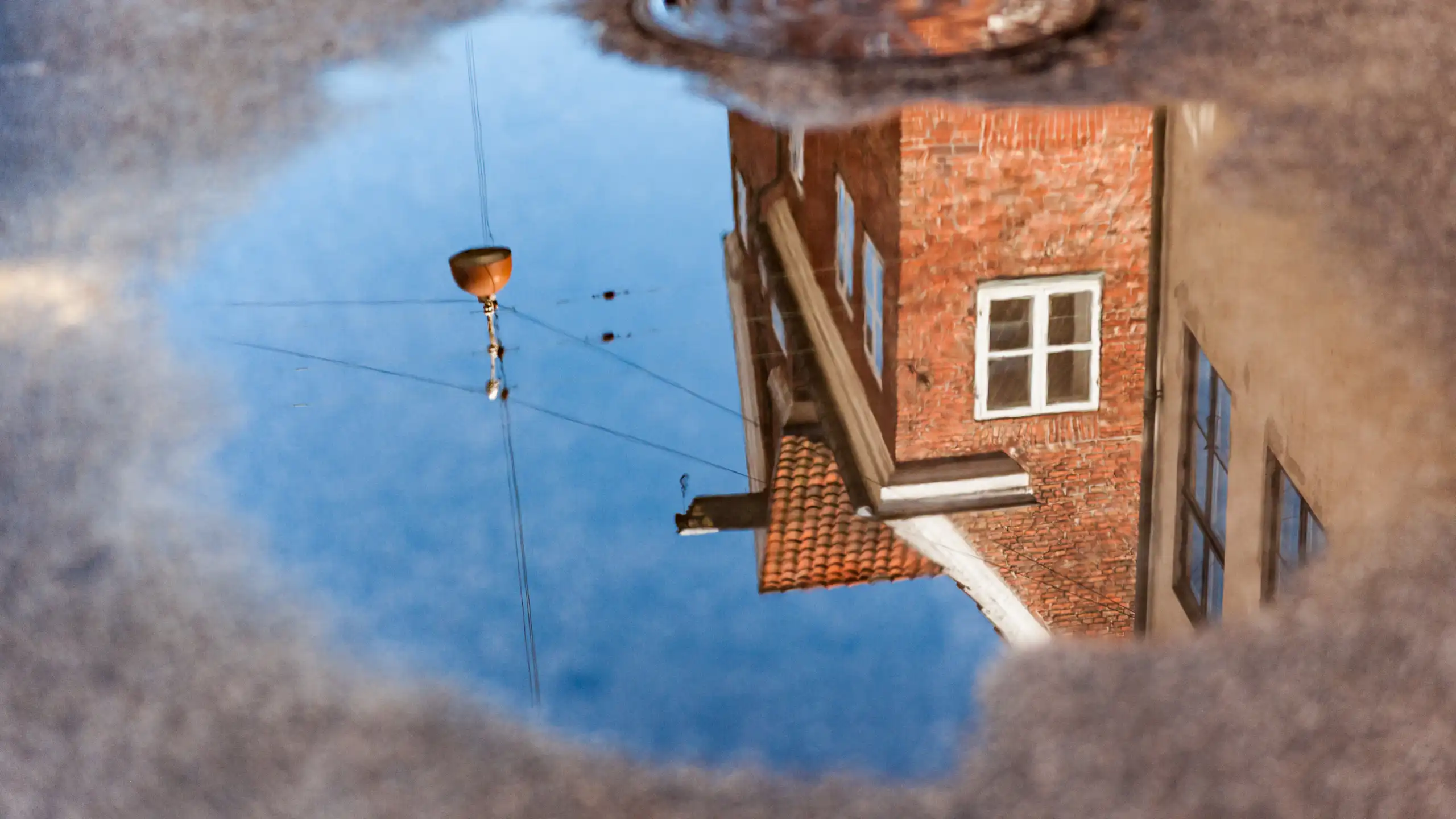 The reflection of a brick house is seen in a small puddle on the road.