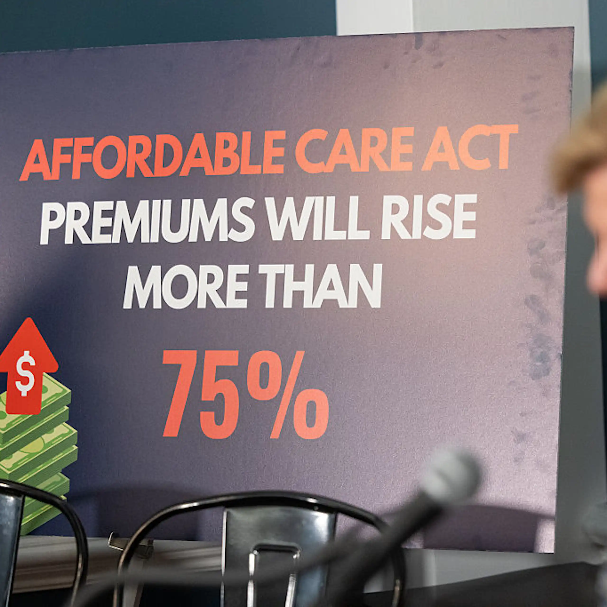 woman standing at a microphone in front of a sign that reads, Affordable Care Act premiums will rise more than 75%