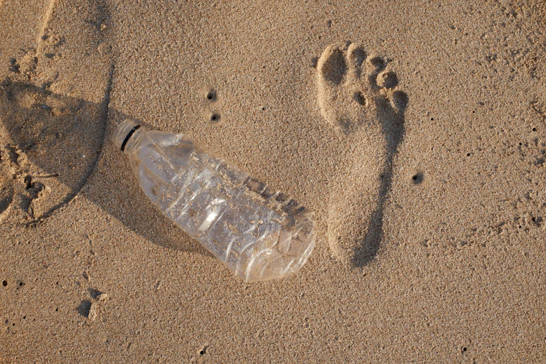 Une empreinte de pas sur le sable de la plage posée à côté d’une bouteille en plastique, vide et chiffonnée.