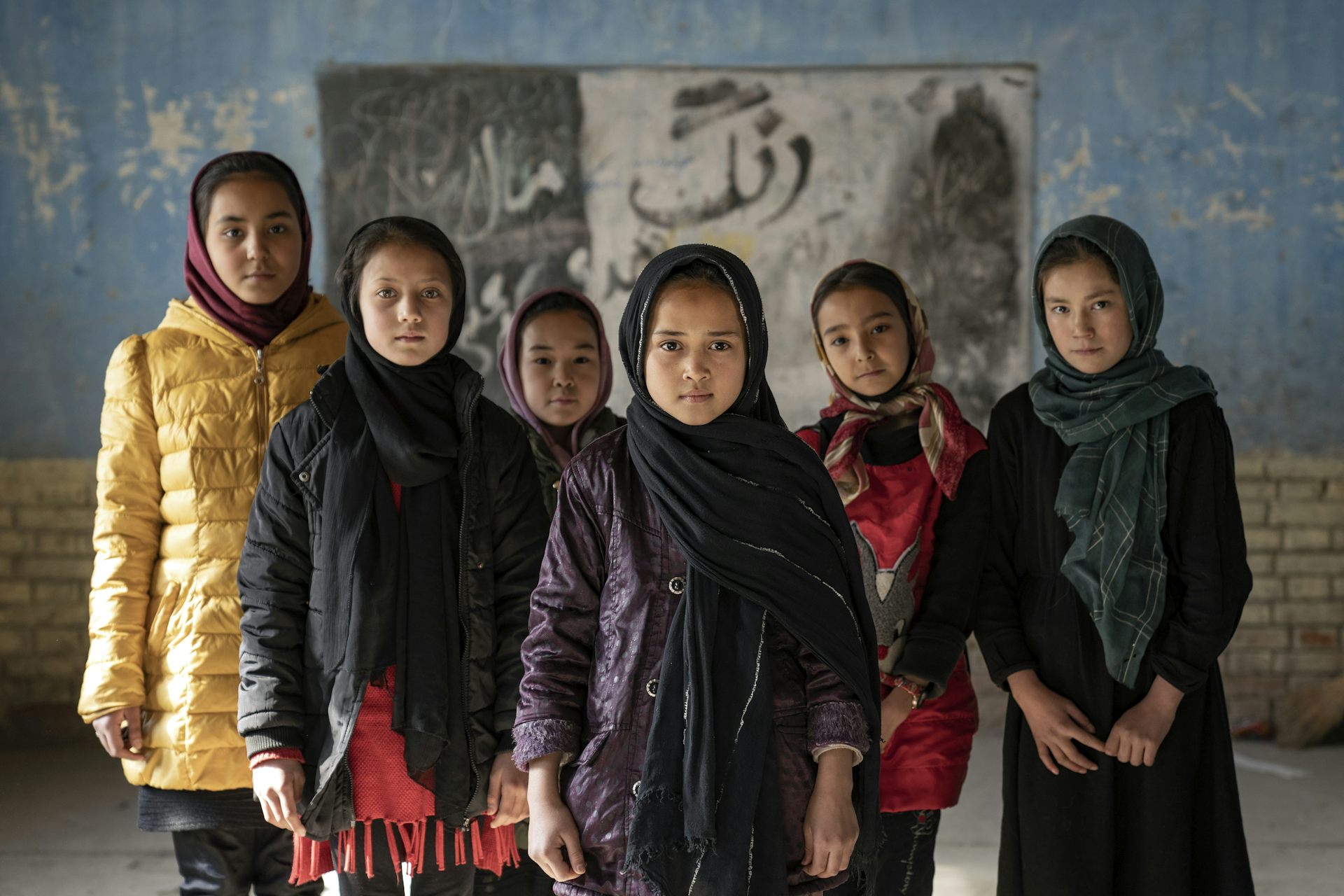 A group of young girls strand shoulder to shoulder facing the camera with solemn looks.