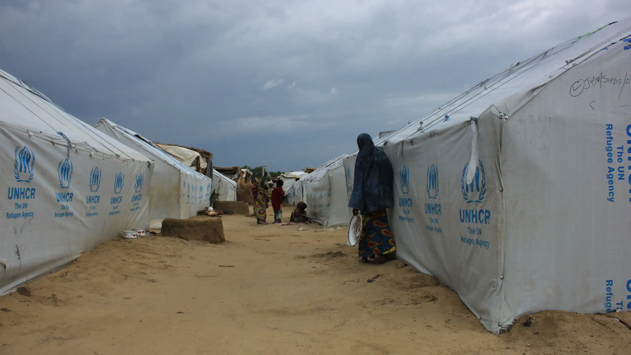 People standing outside white tents marked UNHCR that have been erected on bare ground.