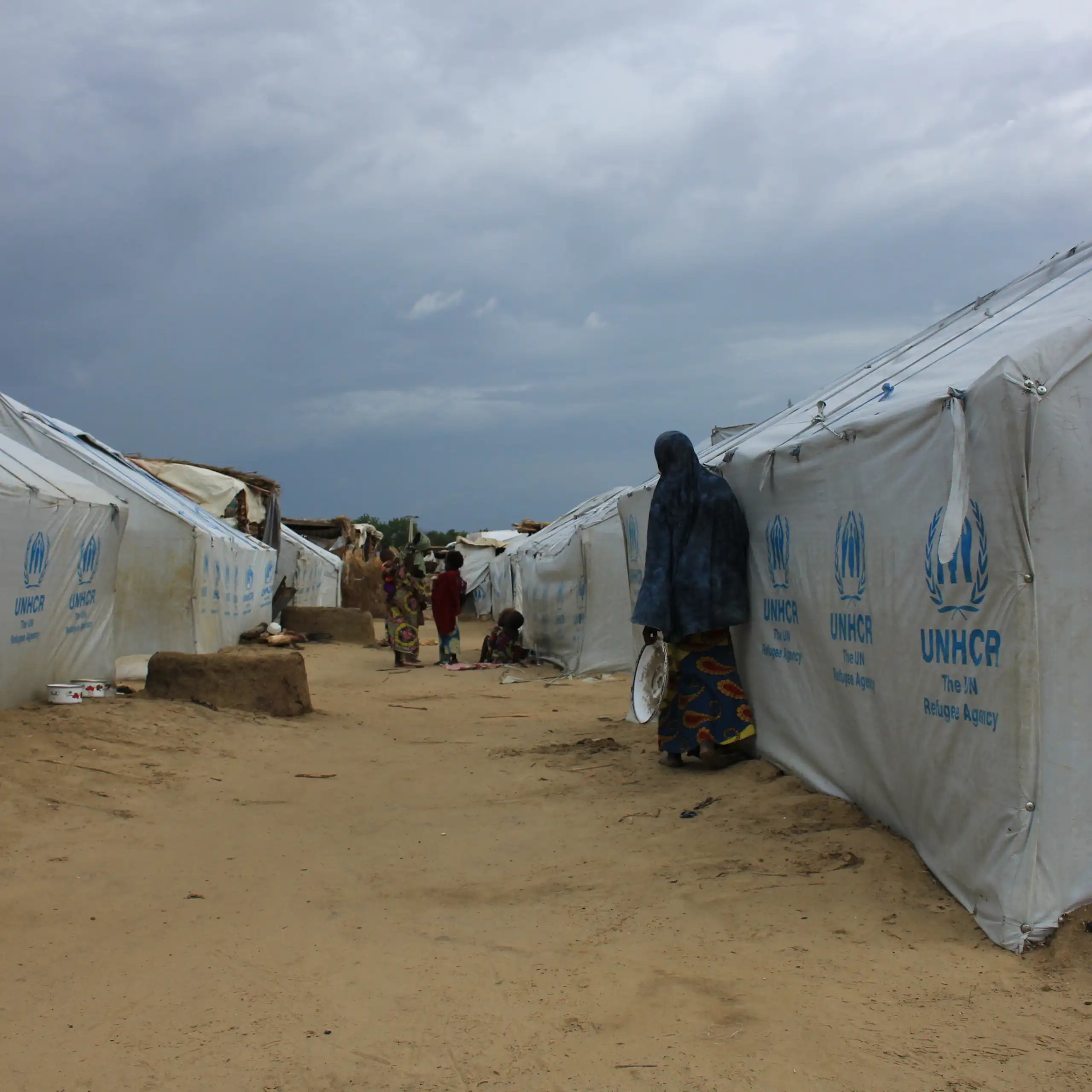 People standing outside white tents marked UNHCR that have been erected on bare ground.