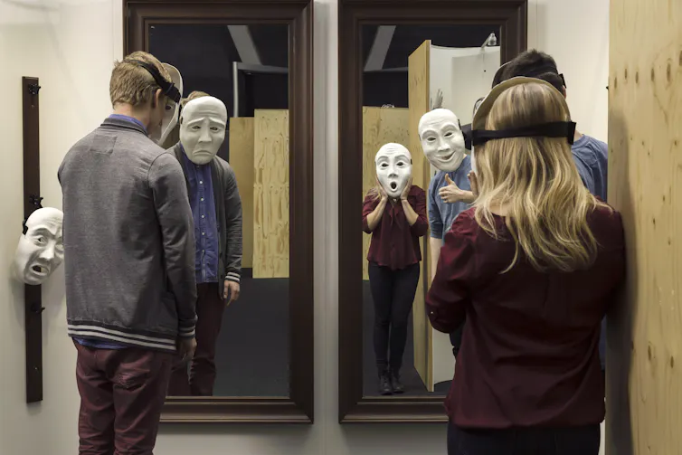 People standing looking in mirrors wearing giant masks.