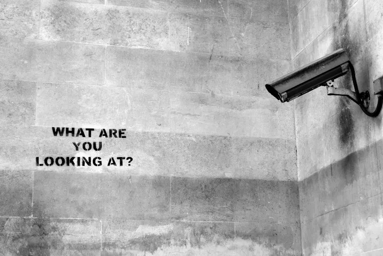 A narrow security camera mounted to a wall points at black lettering that says What Are You Looking At?