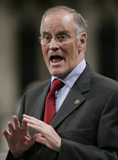 A man in a suit wearing glasses gestures as he speaks.