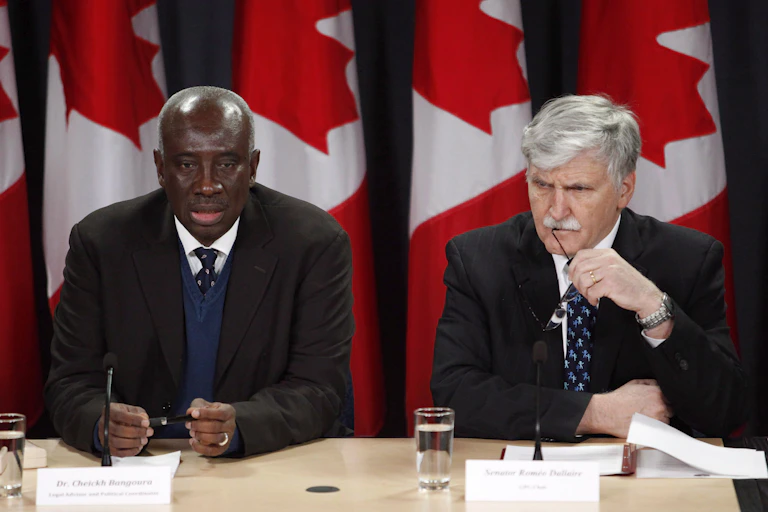 A Black man and a white man with white hair and a white moustache sit at a table in front of a row of Canadian flags.