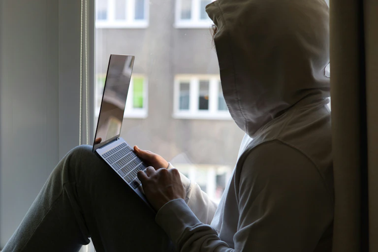 A young person in a hoodie, with a laptop in their hand, looks out of their window.