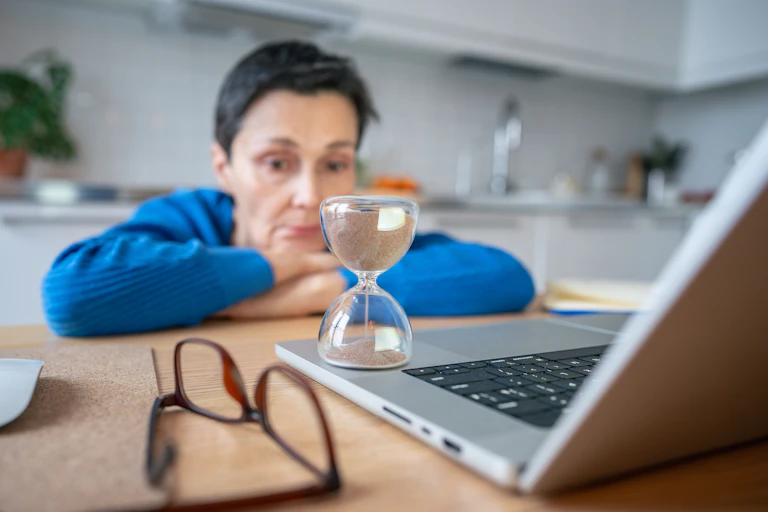A stressed-looking woman rests her head on her hands as she stares at her laptop and an hourglass.
