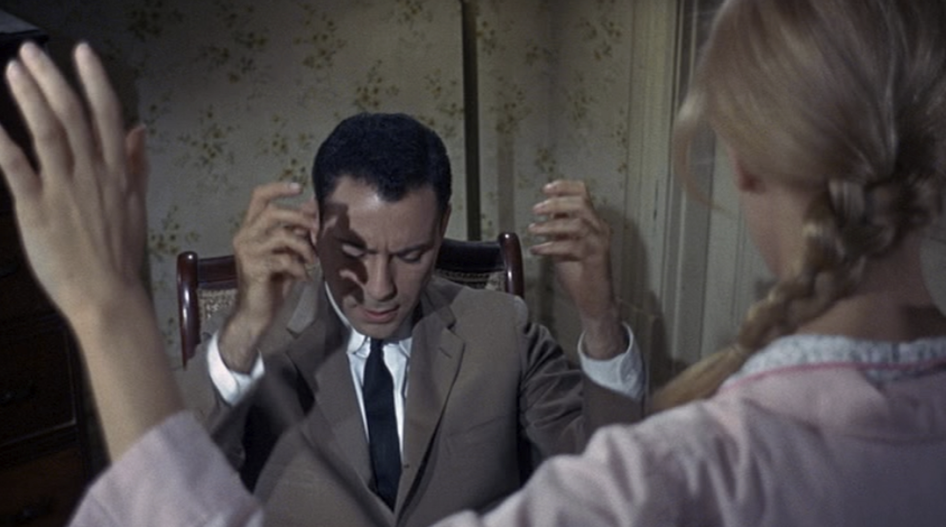 Movie still of a seated man wearing a suit, with his eyes closed, his head down, and his hands raised.
