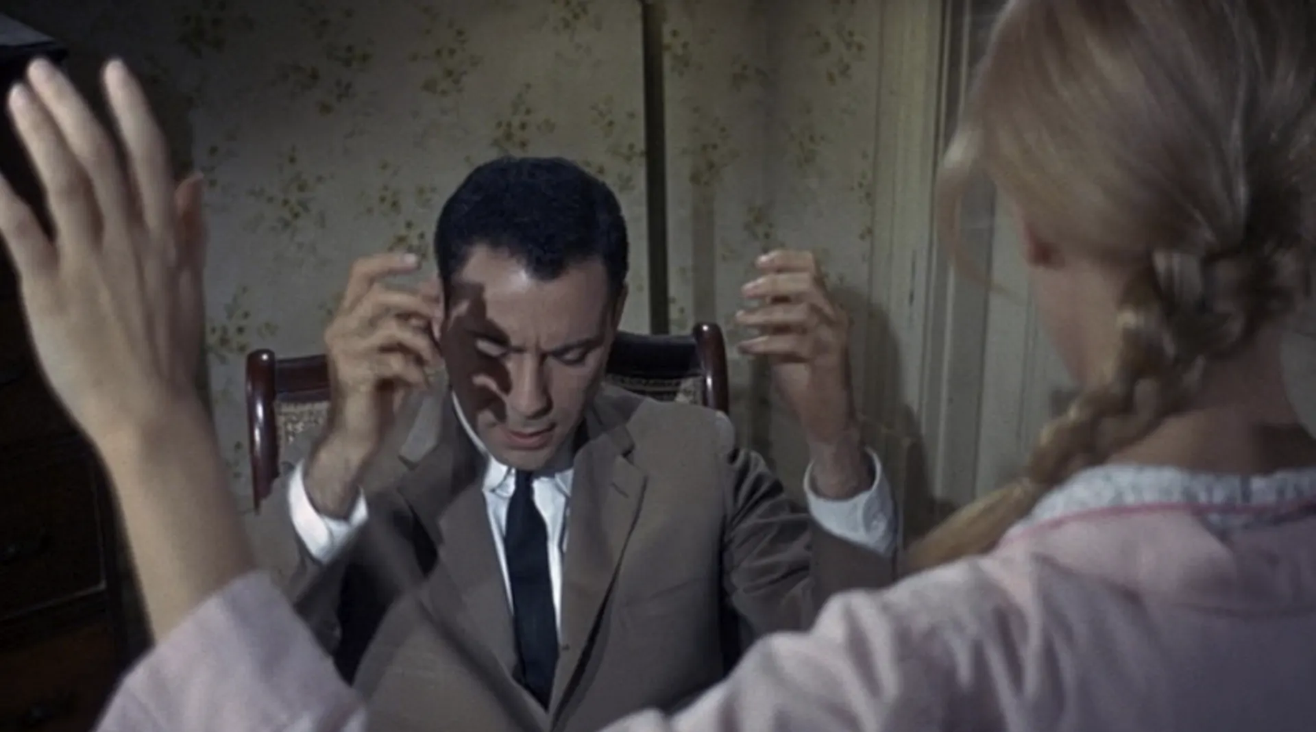 Movie still of a seated man wearing a suit, with his eyes closed, his head down, and his hands raised.