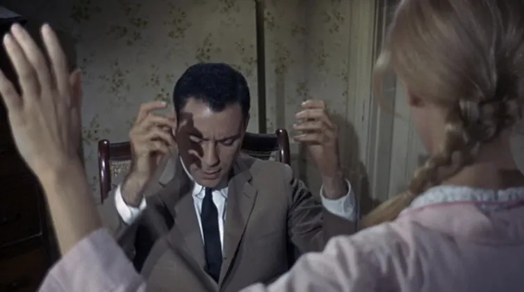 Movie still of a seated man wearing a suit, with his eyes closed, his head down, and his hands raised.