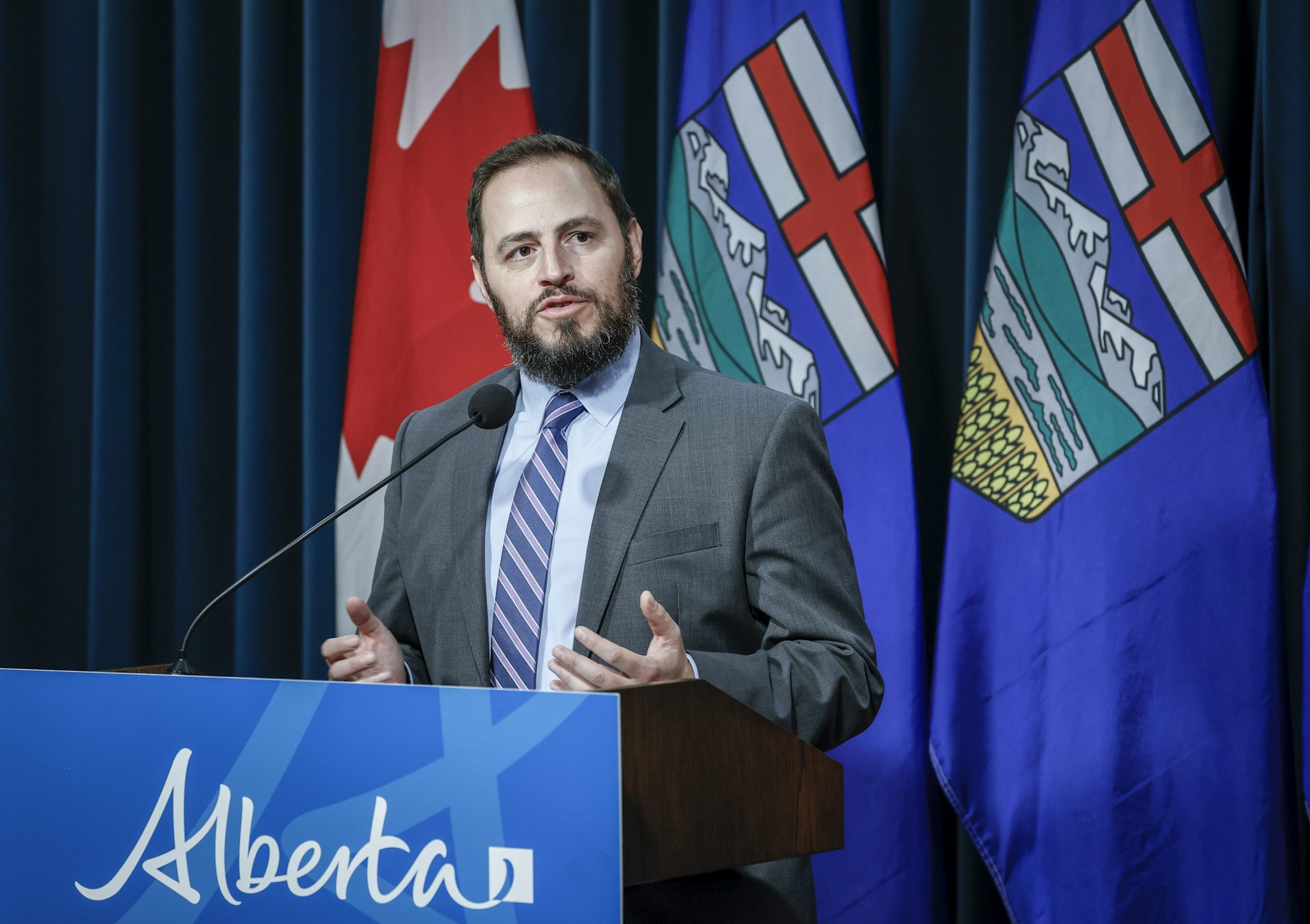 A man in a suit at a podium with a sign that says Alberta and flags behind him.