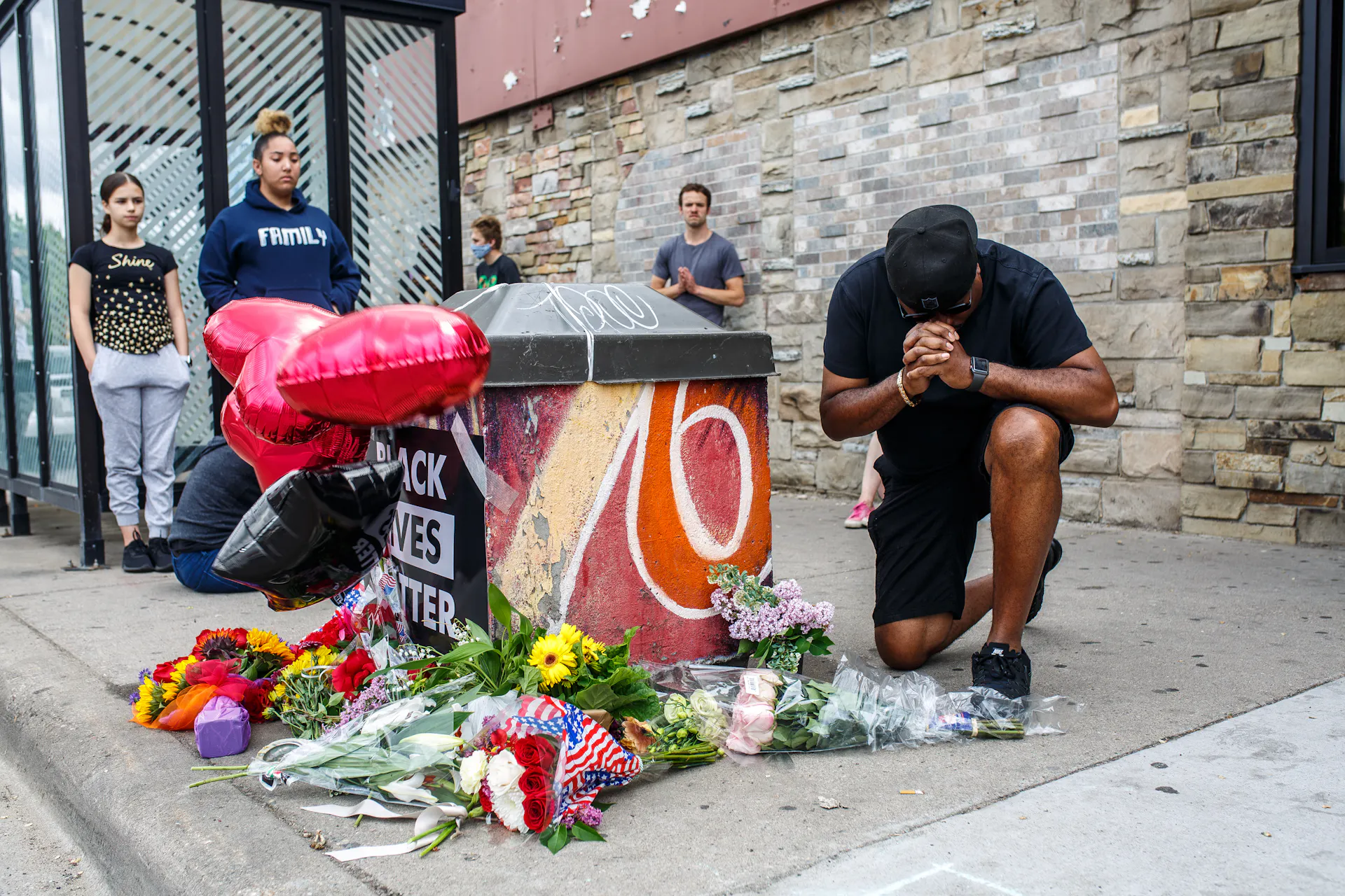 A man kneels in front of a makeshift memorial next to a bus stop on a city street.