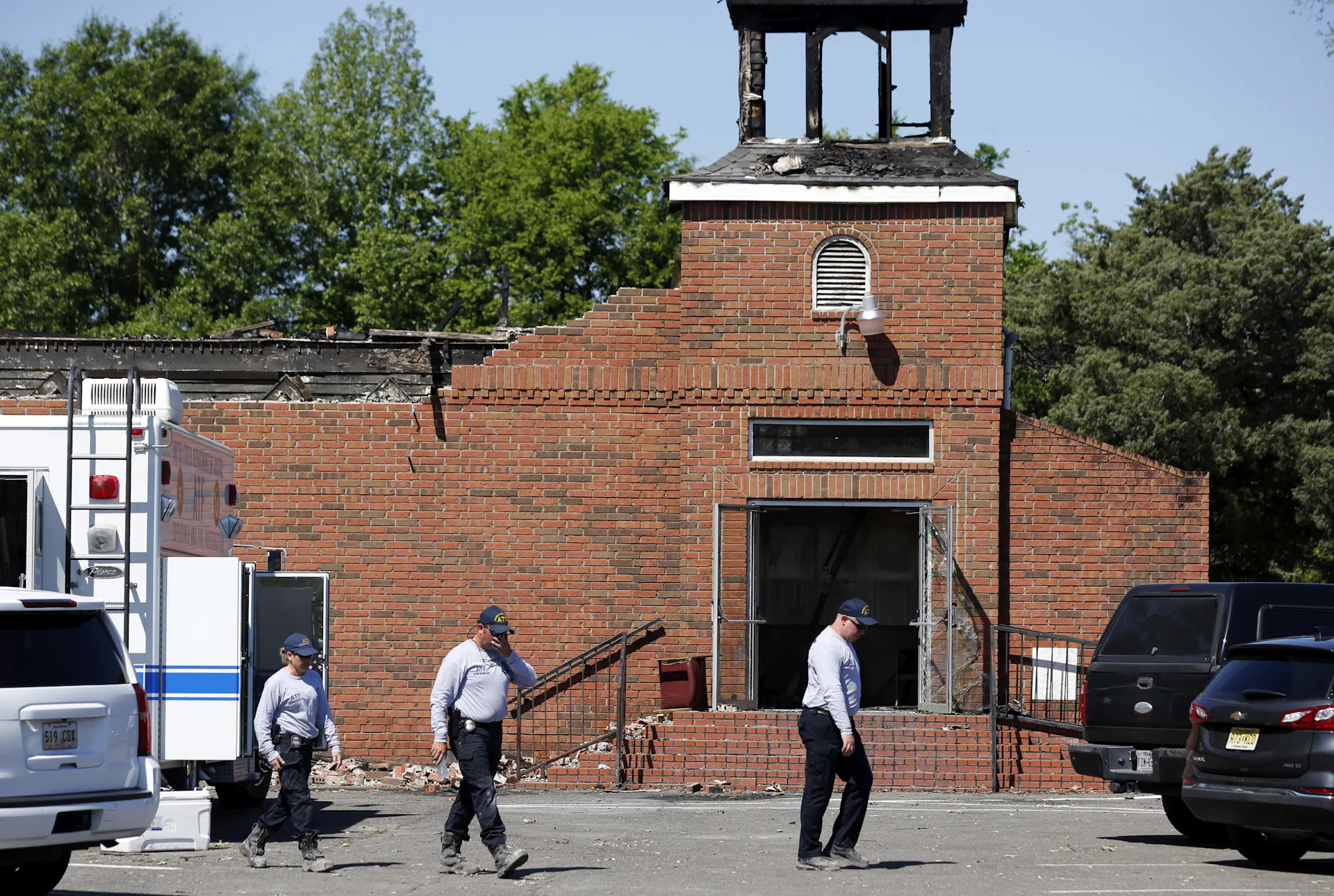 Three people in white shirts and dark pants walk across a parking lot outside a destroyed brick building.