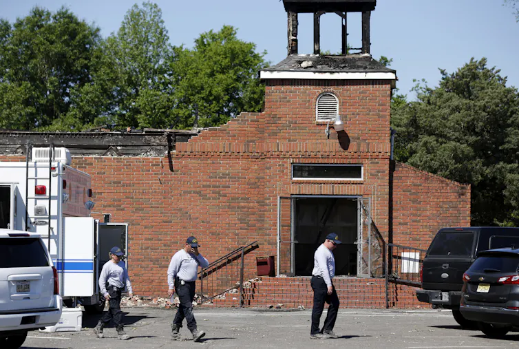 Three people in white shirts and dark pants walk across a parking lot outside a destroyed brick building.