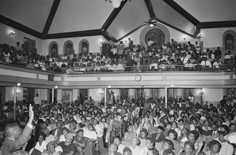 A black and white photo of a crowded church, with people packing the pews of the main sanctuary and balcony.