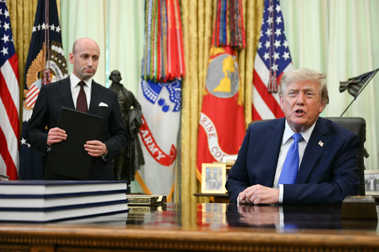 A man seated at a desk speaks as another many standing in the background looks at him.