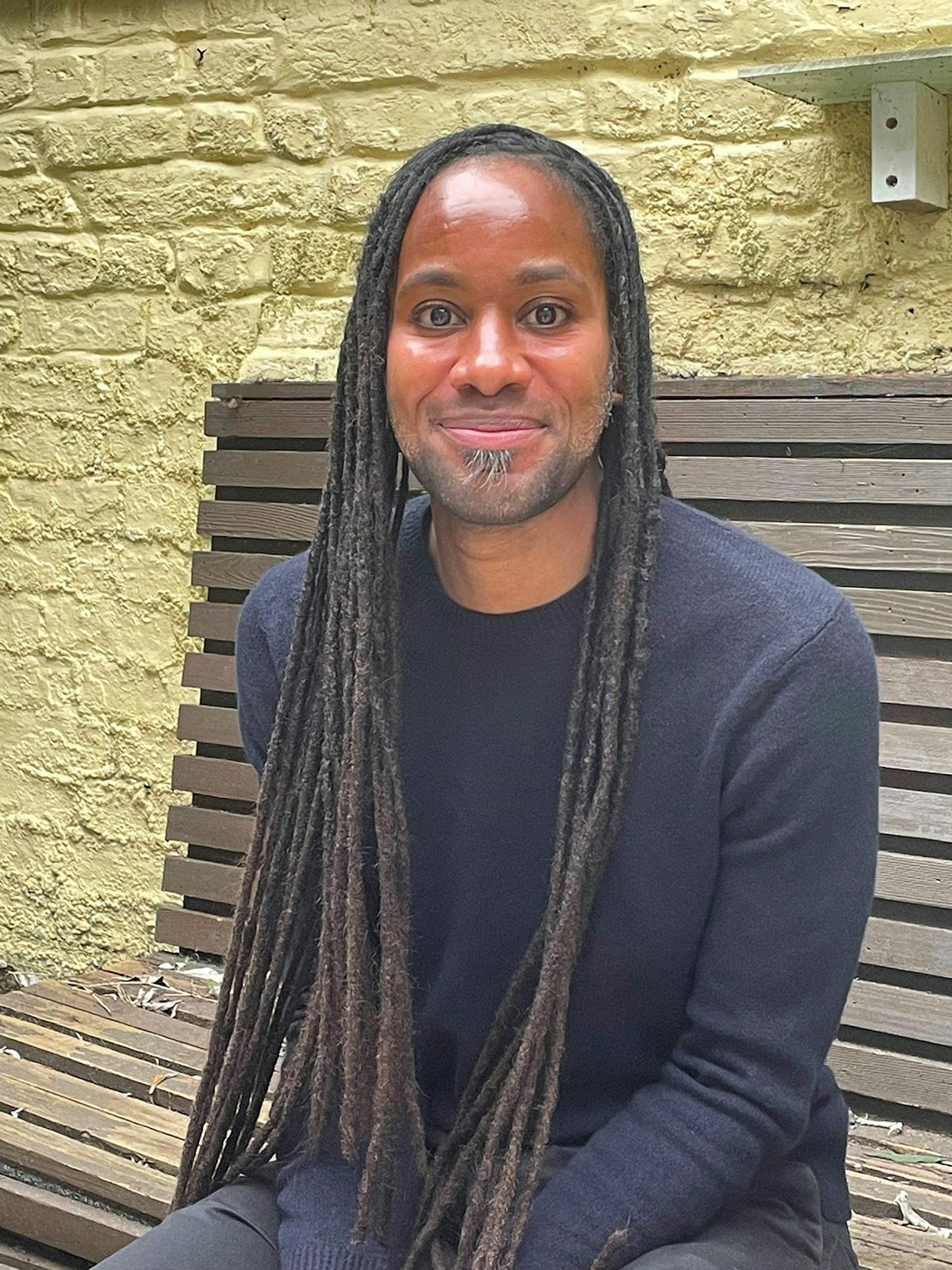 A photo of a smiling man, (no teeth showing), long black locks, blue sweater sitting on a bench
