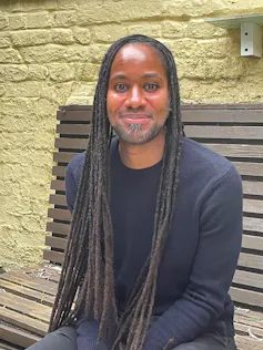 A photo of a smiling man, (no teeth showing), long black locks, blue sweater sitting on a bench