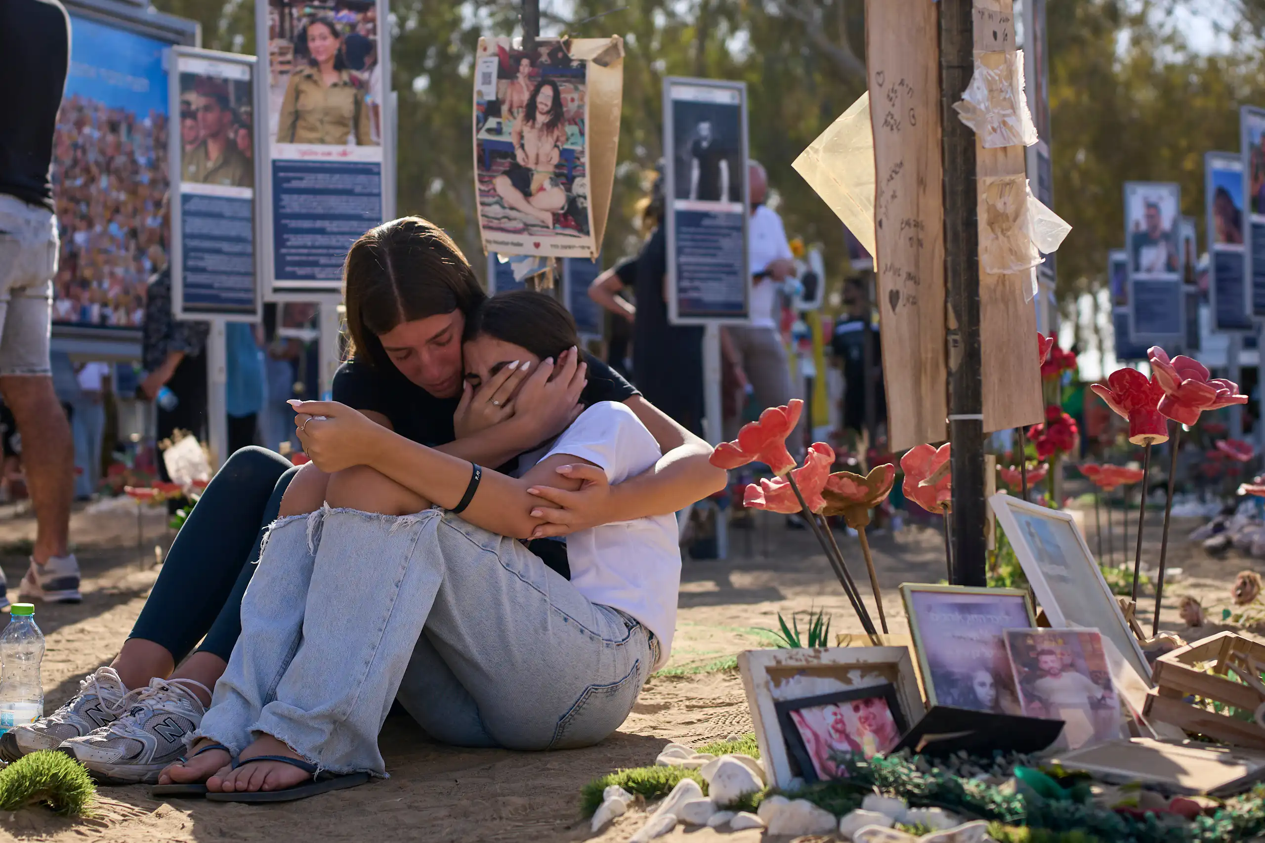 Two women sitting on the floor and crying as they hug each other at a remembrance site.
