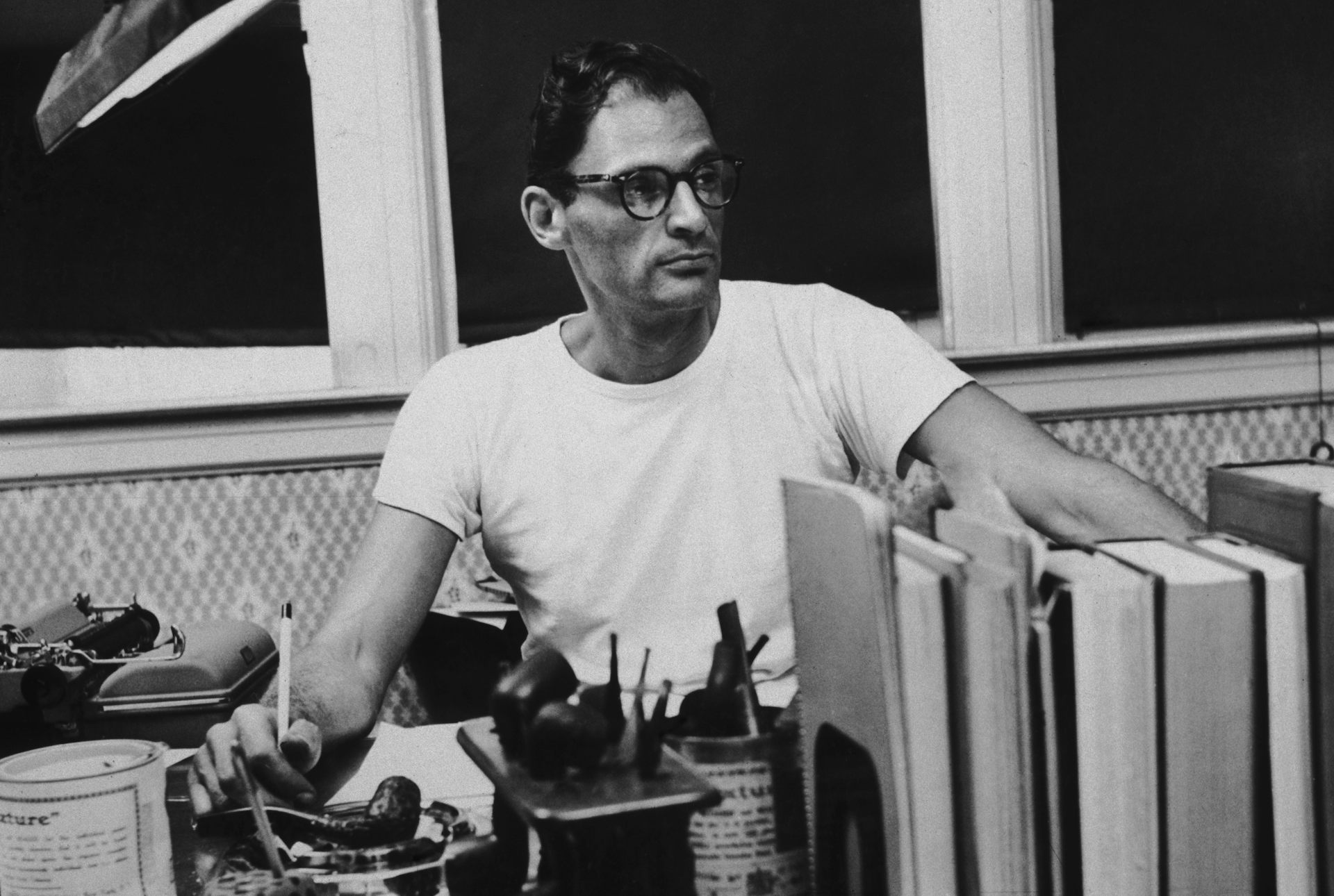 Black and white image, Miller holds a pen, lots of books present.