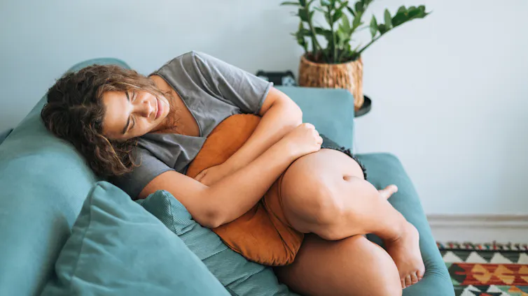 Young woman in pain sitting on sofa, clutching pillow to tummy