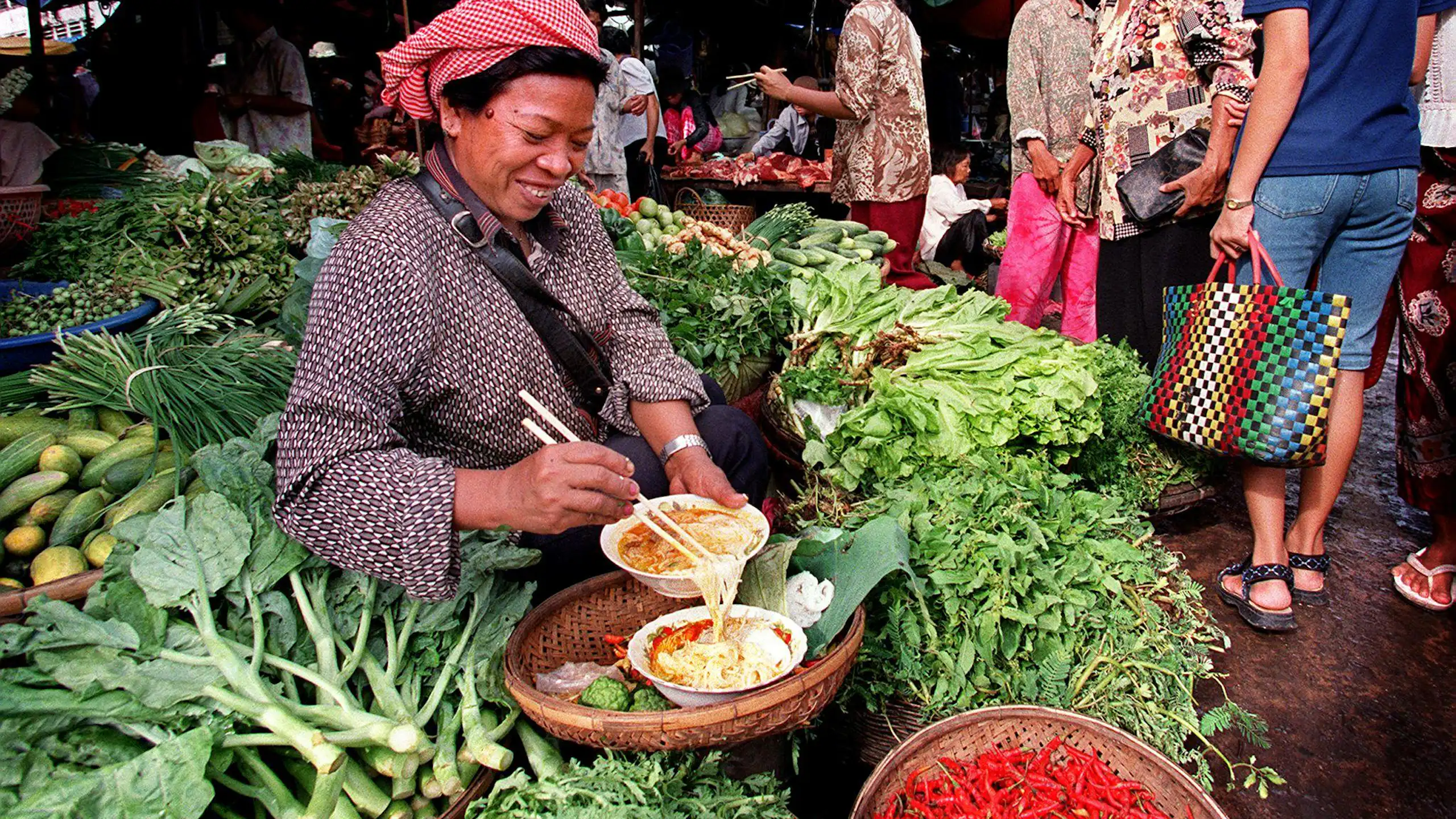 Shoppers pass by a Cambodian vendor sitting amongst a colourful bed of vegetables.
