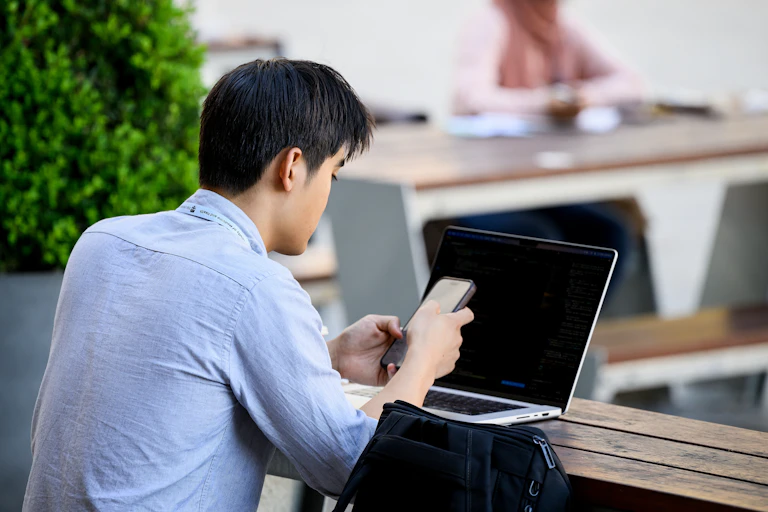 A male university student using his phone and laptop, sitting at a bench outside. A female student is studying at another table nearby.