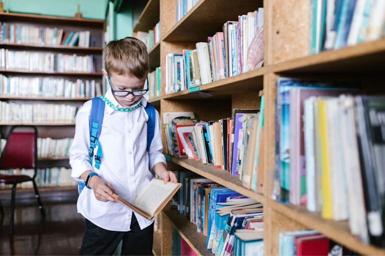A young boy in a backpack reads a book in a library.