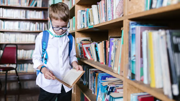 A young boy in a backpack reads a book in a library.