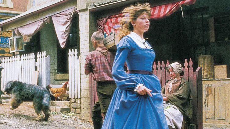 A young woman in an old-fashioned dress walking fast past old houses in a narrow-looking street. A dog in background.