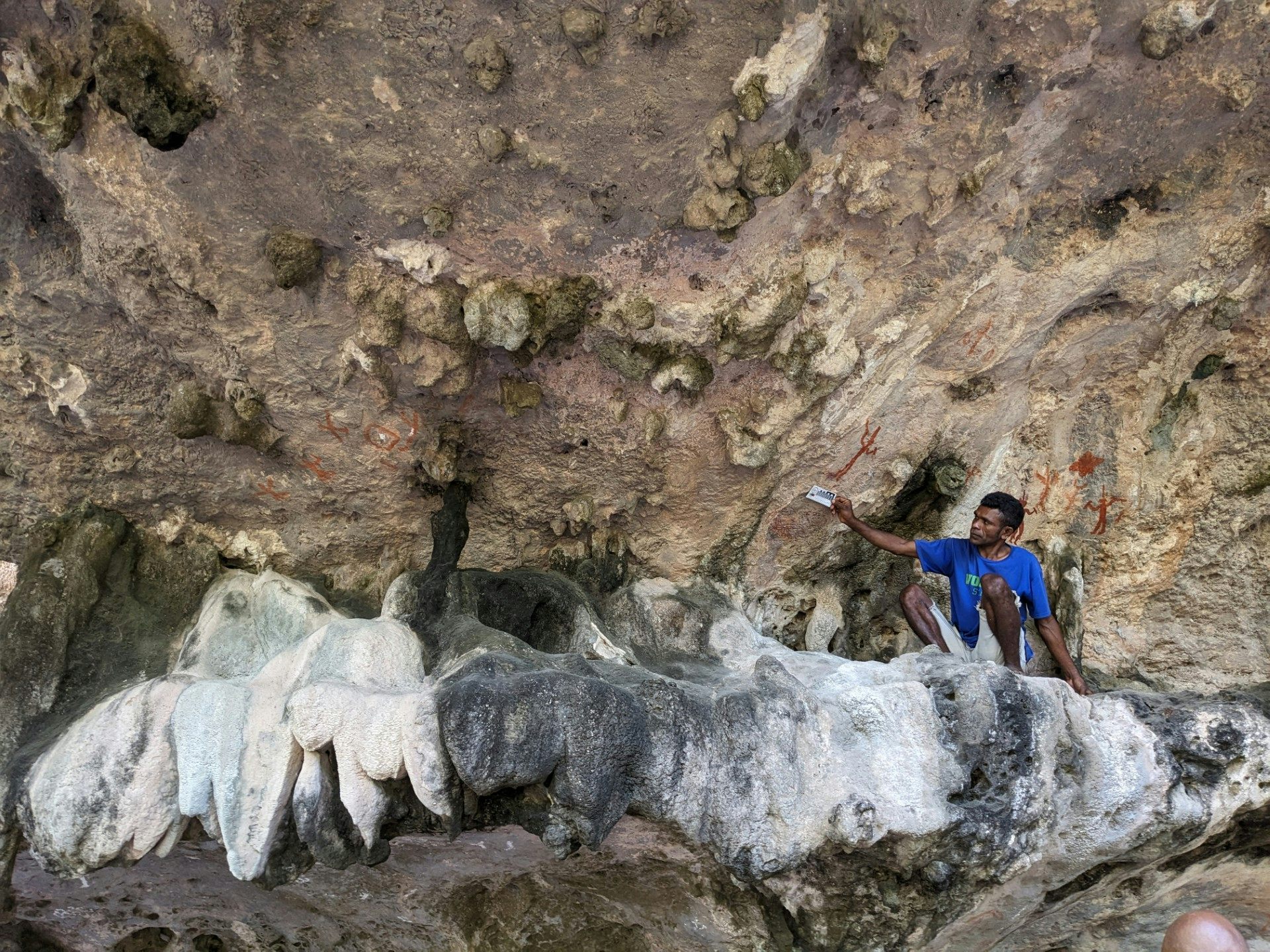 A man sits underneath what looks like a rocky outcrop, with red pigment visible on the walls behind.