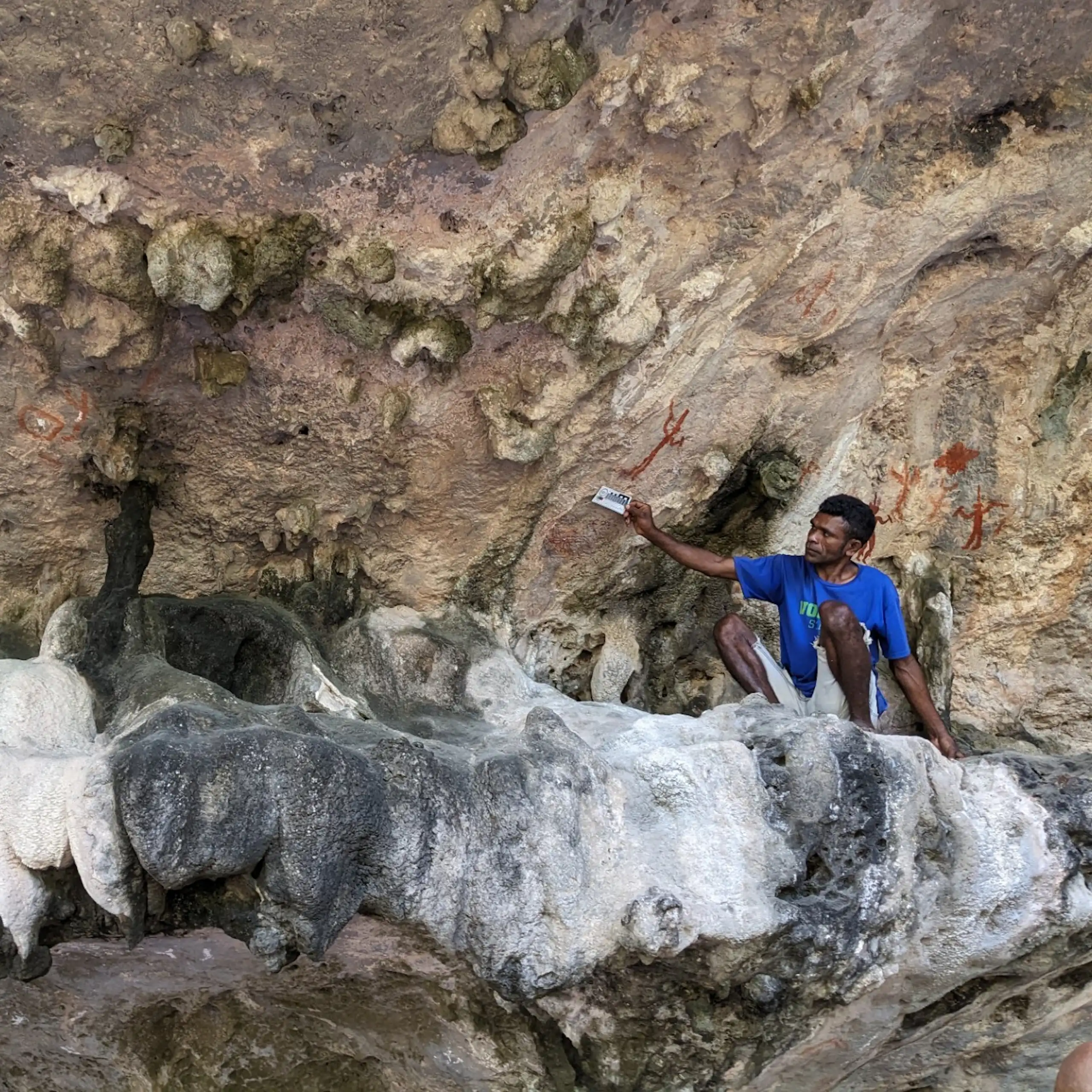 A man sits underneath what looks like a rocky outcrop, with red pigment visible on the walls behind.