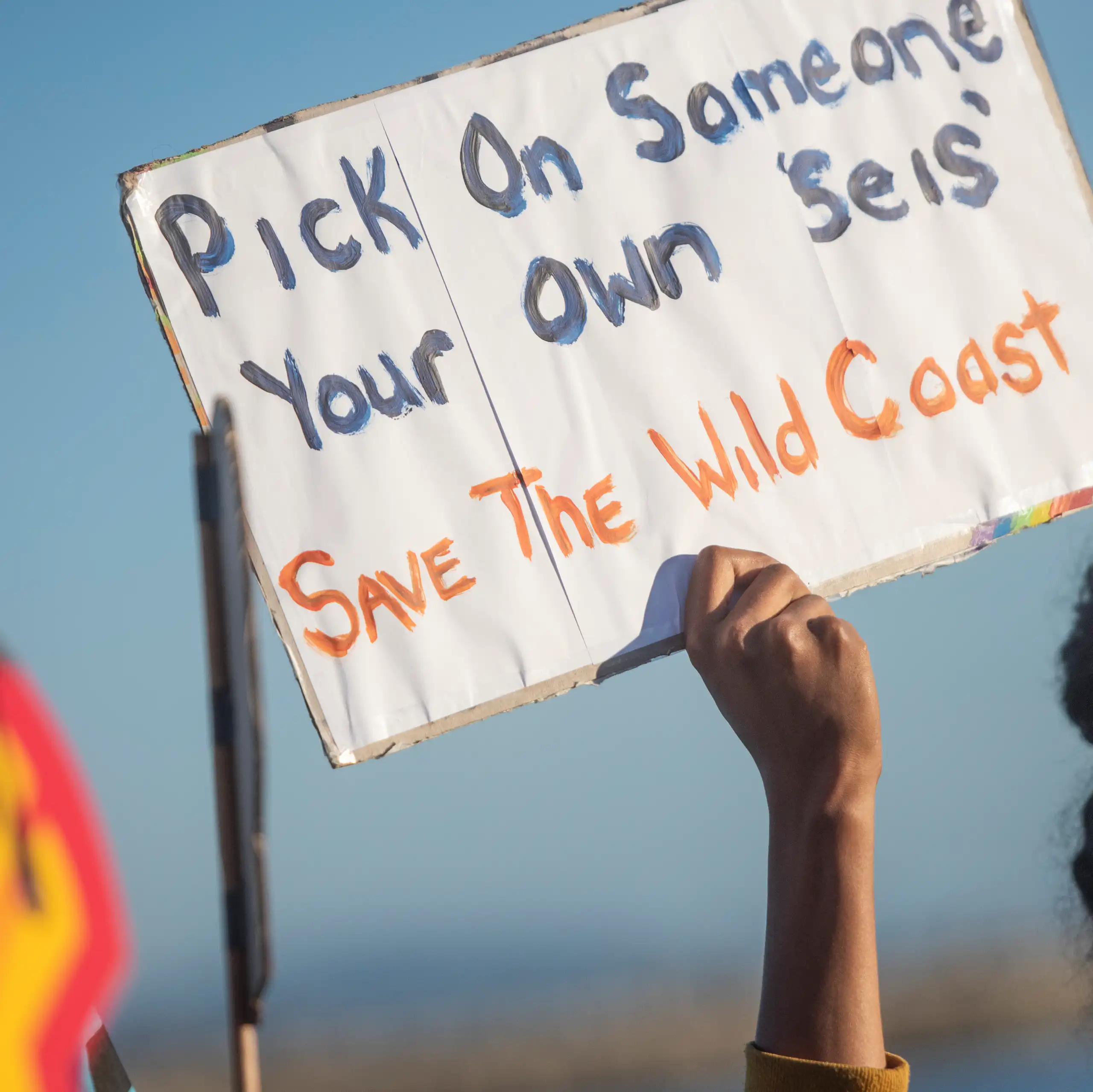 Person holds up placard saying 'Pick on someone your own seis, Save the Wild Coast' and another person holds a giant Shell logo that shows oil dripping down like blood