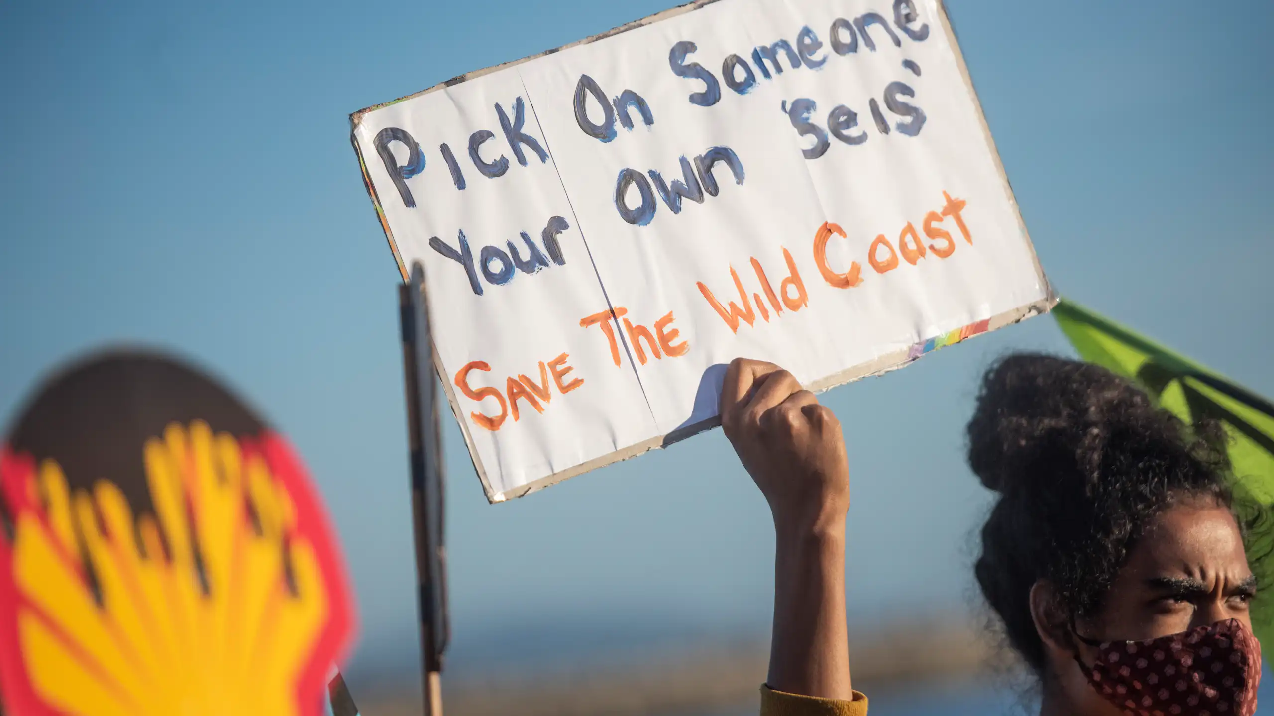 Person holds up placard saying 'Pick on someone your own seis, Save the Wild Coast' and another person holds a giant Shell logo that shows oil dripping down like blood