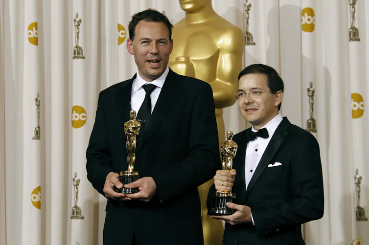 Two suited men standing in front of a curtain and a giant gold coloured Oscar statue, each holding an Oscar trophy.