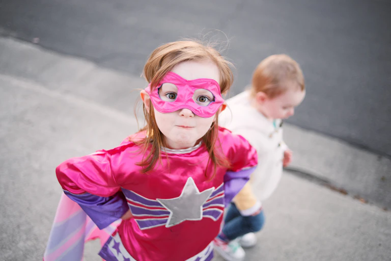 Young girl wearing superhero costume looks up at the camera while a younger child out of costume is seen behind her.